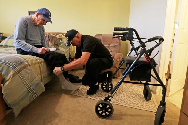 Caregiver Warren Manchess helping Paul Gregoline with his shoes and socks, in Noblesville, Ind.